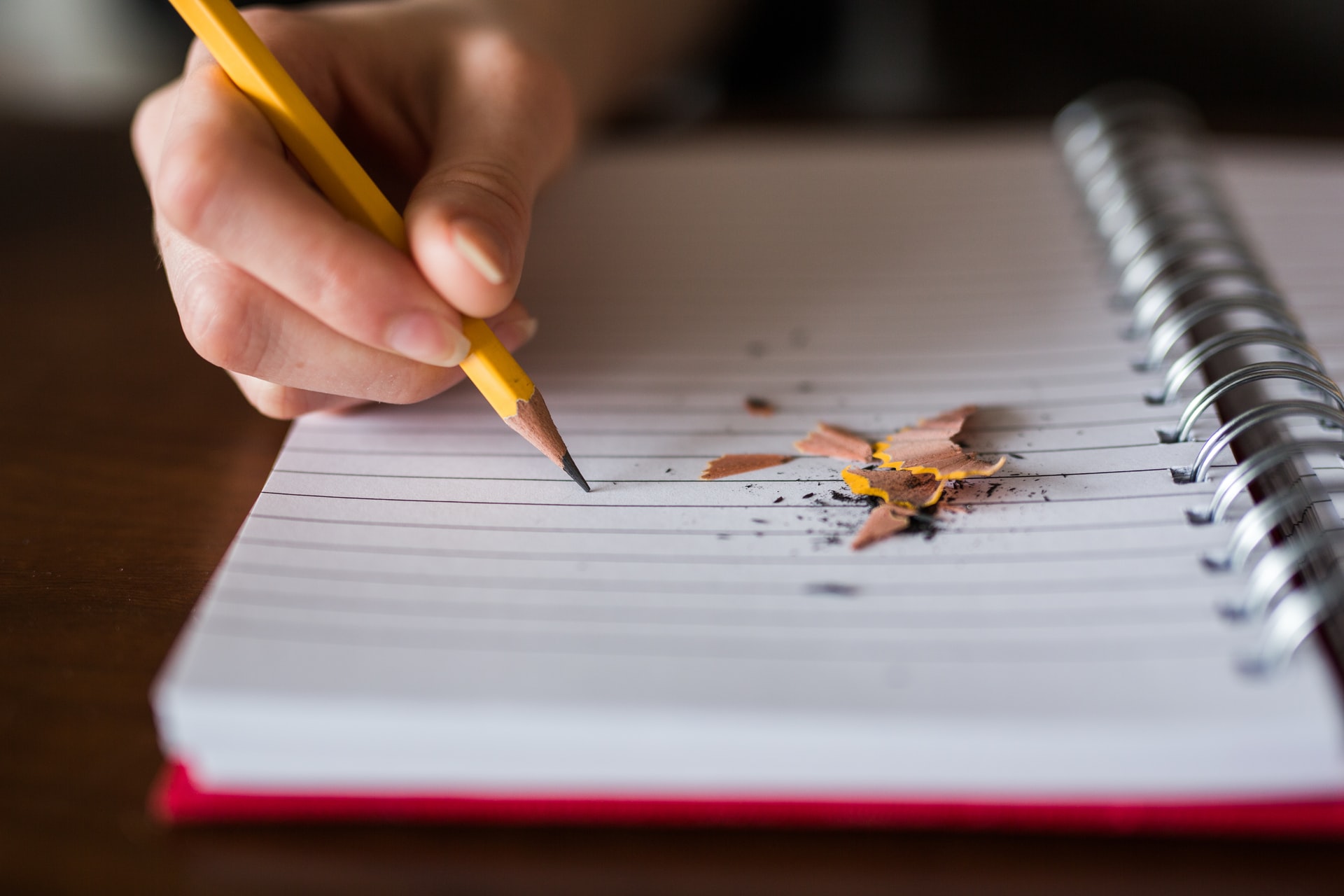 An image of a blank notebook and someone holding a pencil to the paper, pencil shavings on the paper